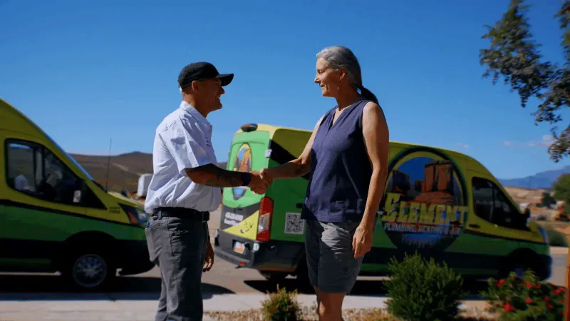 Element plumbing heating air technician shaking hands with a customer in st george ut in front of branded service vans with a scenic mountain backdrop in St. George, UT