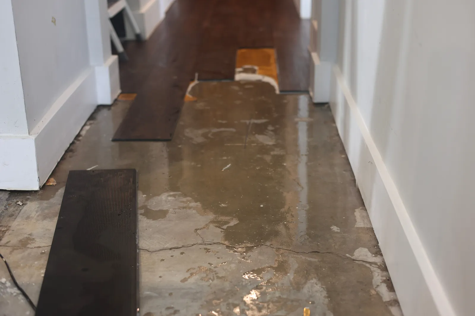 Water damage from a foundation slab leak: flooring pulled up and standing water on a cracked concrete slab in a hallway
